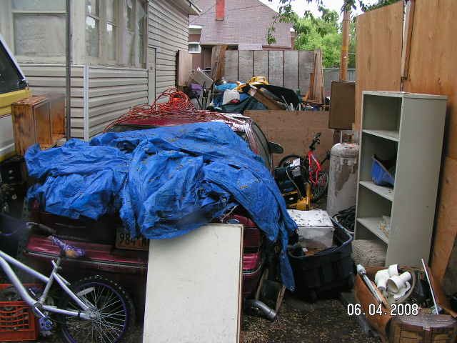A junk filled yard with a car with a blue tarp over it.